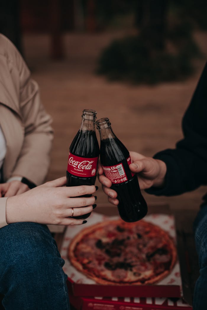 Two people toast with Coca-Cola bottles over a pizza. Warm, social setting.